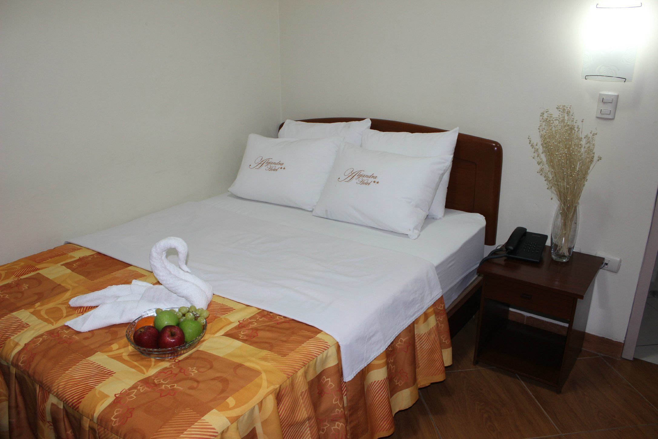 Una cama bien hecha con sábanas blancas, un frutero sobre la cama y un florero con flores secas en la mesita de noche.