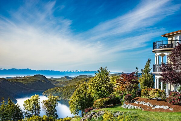 A scenic view of a lakeside property with a prominent building featuring a balcony and a well-maintained garden. - Villa Eyrie Resort (Malahat)