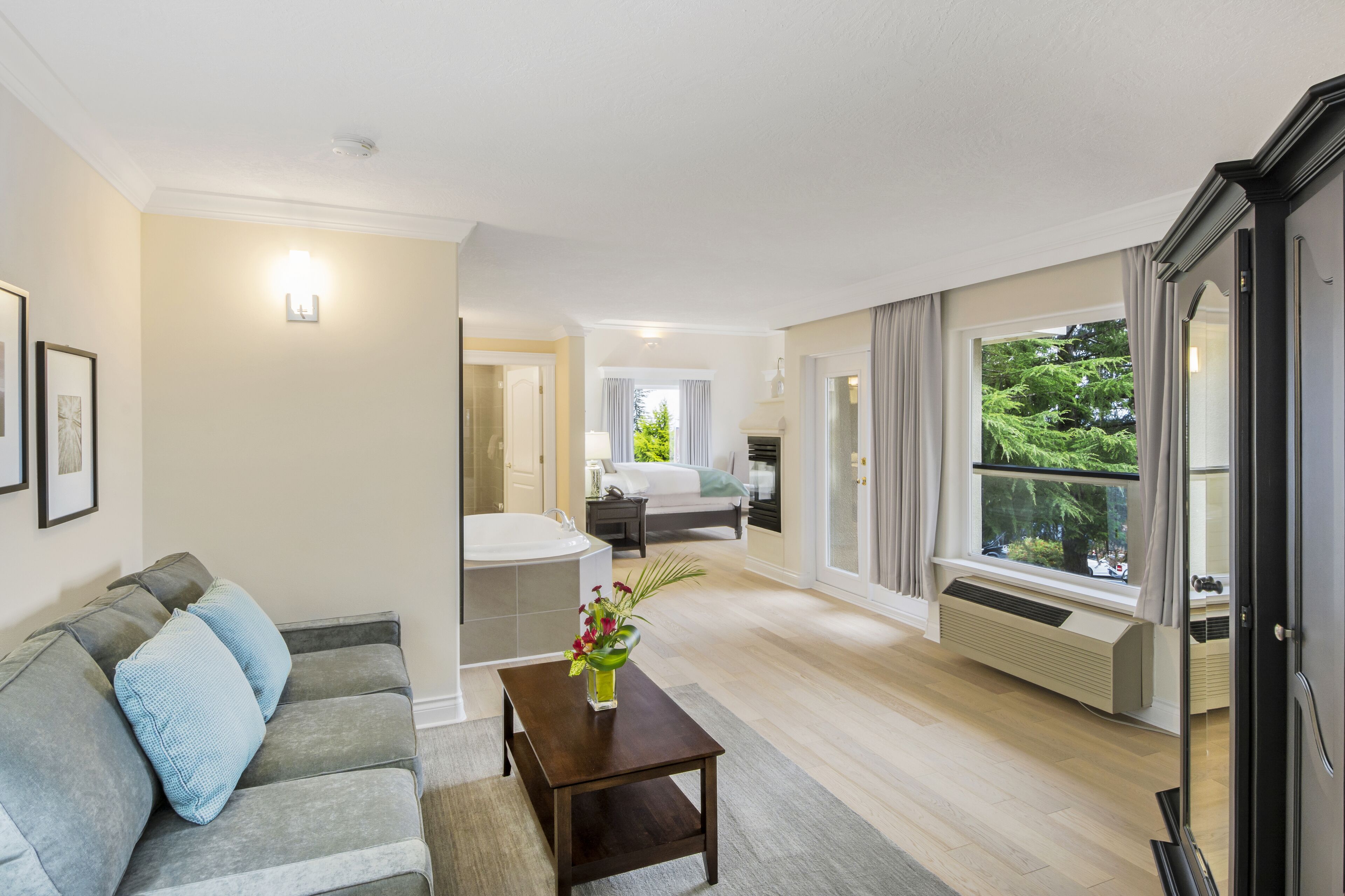 Una sala moderna con un sofá gris, una mesita de madera con un florero, un ventanal con cortinas y vistas al exterior con vegetación.