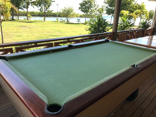 Une table de billard sur une terrasse en bois, avec vue sur une zone herbeuse et des arbres.