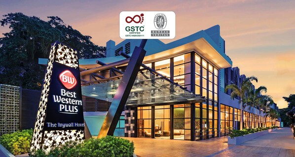 Un hotel moderno con fachada de vidrio, un llamativo letrero de 'Best Western Plus' y certificaciones de GSTC y BUREAU VERITAS.