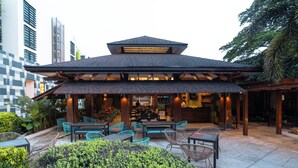 An outdoor cafe with tables and chairs, surrounded by greenery and a building with a sloped roof.