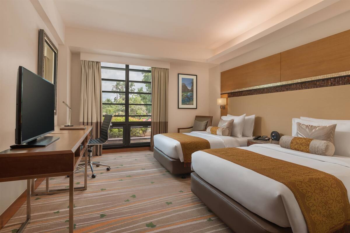 Ein Hotelzimmer mit zwei Betten, einem Schreibtisch mit Fernseher, einem Fenster mit Vorhängen und Blick ins Grüne.
