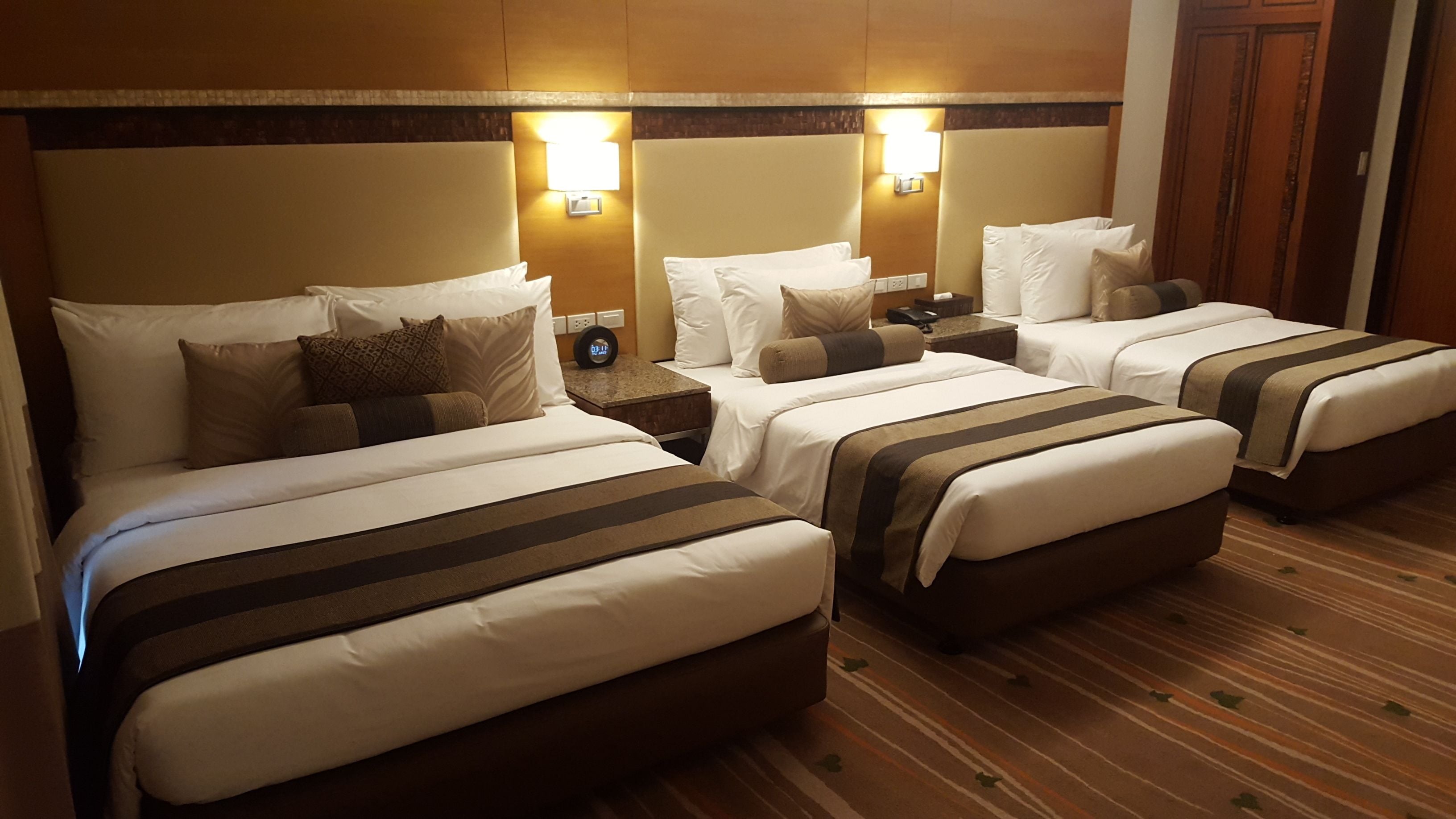 A hotel room with three beds, each with white bedding and brown stripes, a wooden headboard, and a nightstand with a lamp.