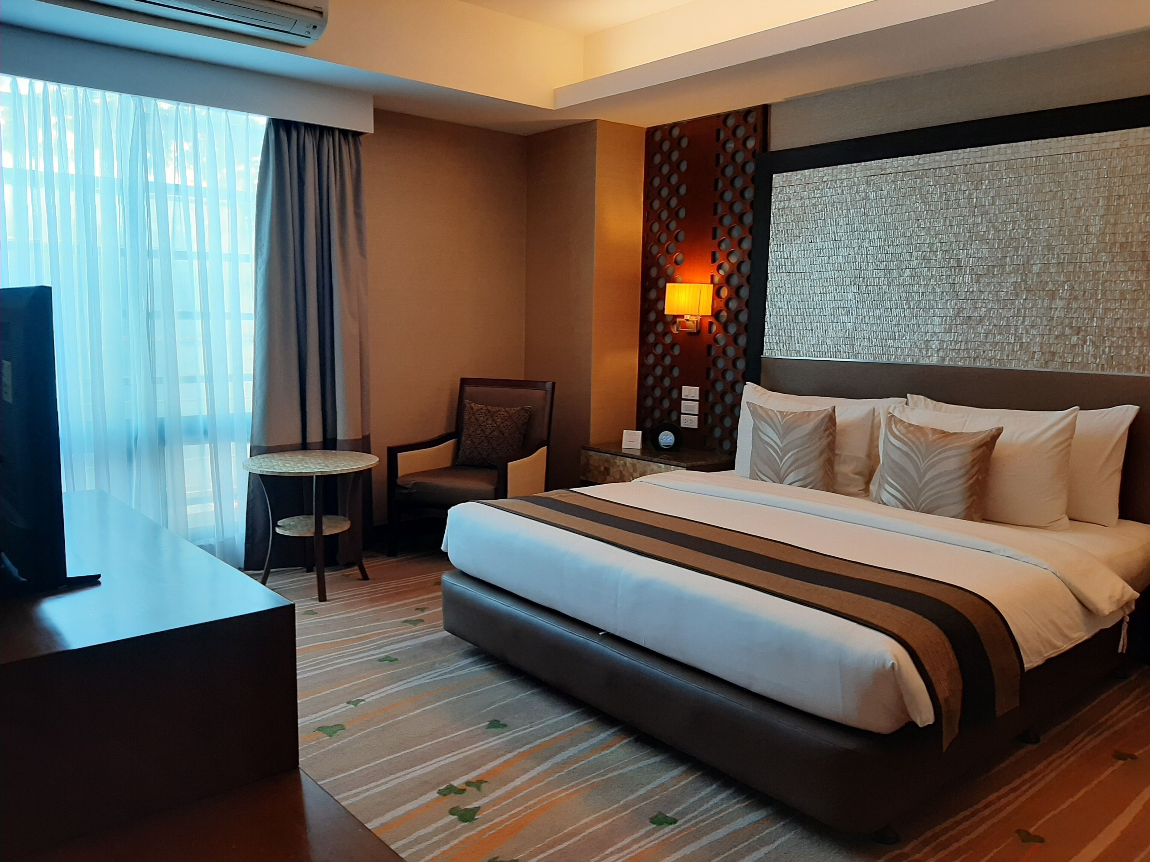 A hotel room with a large bed, a desk, a chair, a small table, and a wall-mounted TV.