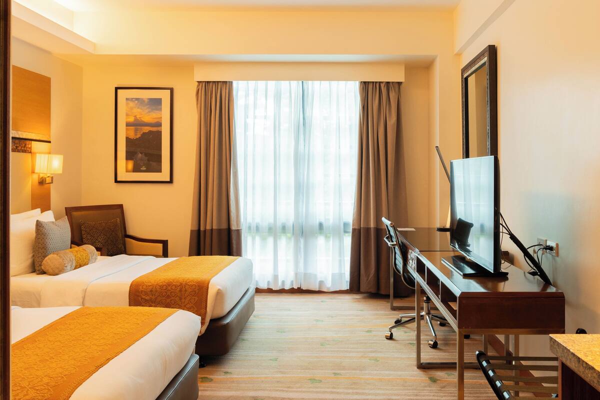 Ein Hotelzimmer mit zwei Betten, einem Schreibtisch mit Fernseher, einem Sessel und einem Fenster mit Vorhängen.