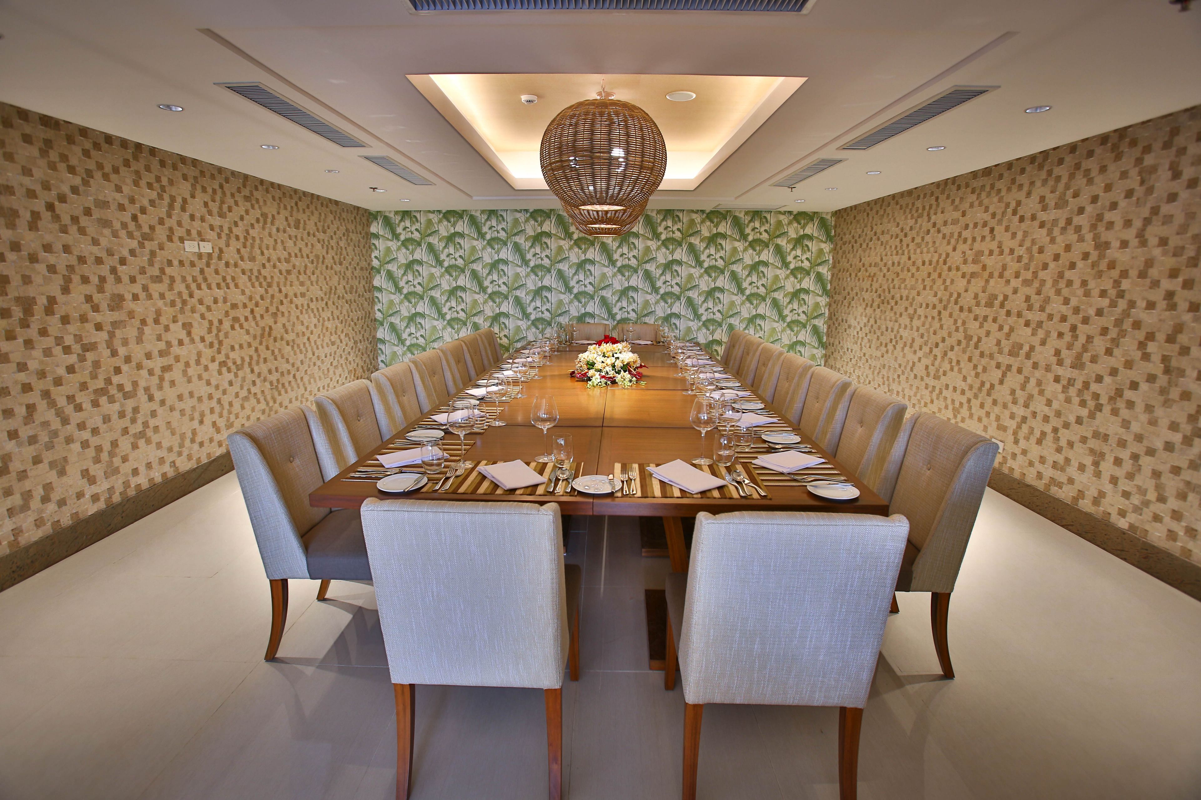 Una larga mesa de comedor dispuesta para una comida formal en una habitación bien iluminada, con papel tapiz estampado y una lámpara decorativa en el techo.