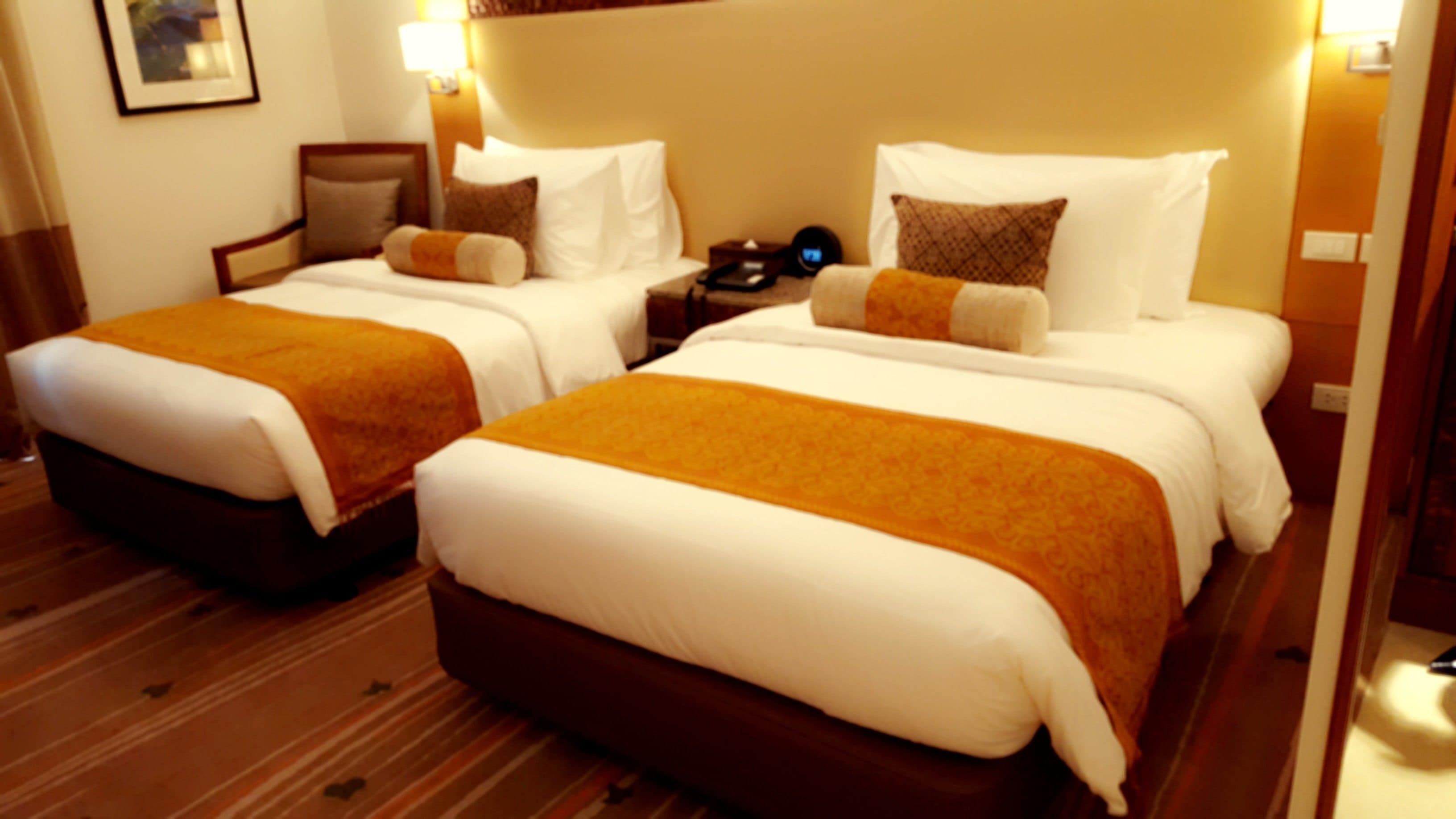 A hotel room with two beds, each with white linens and brown headboards. There are bedside tables with lamps and a clock on one of the tables.