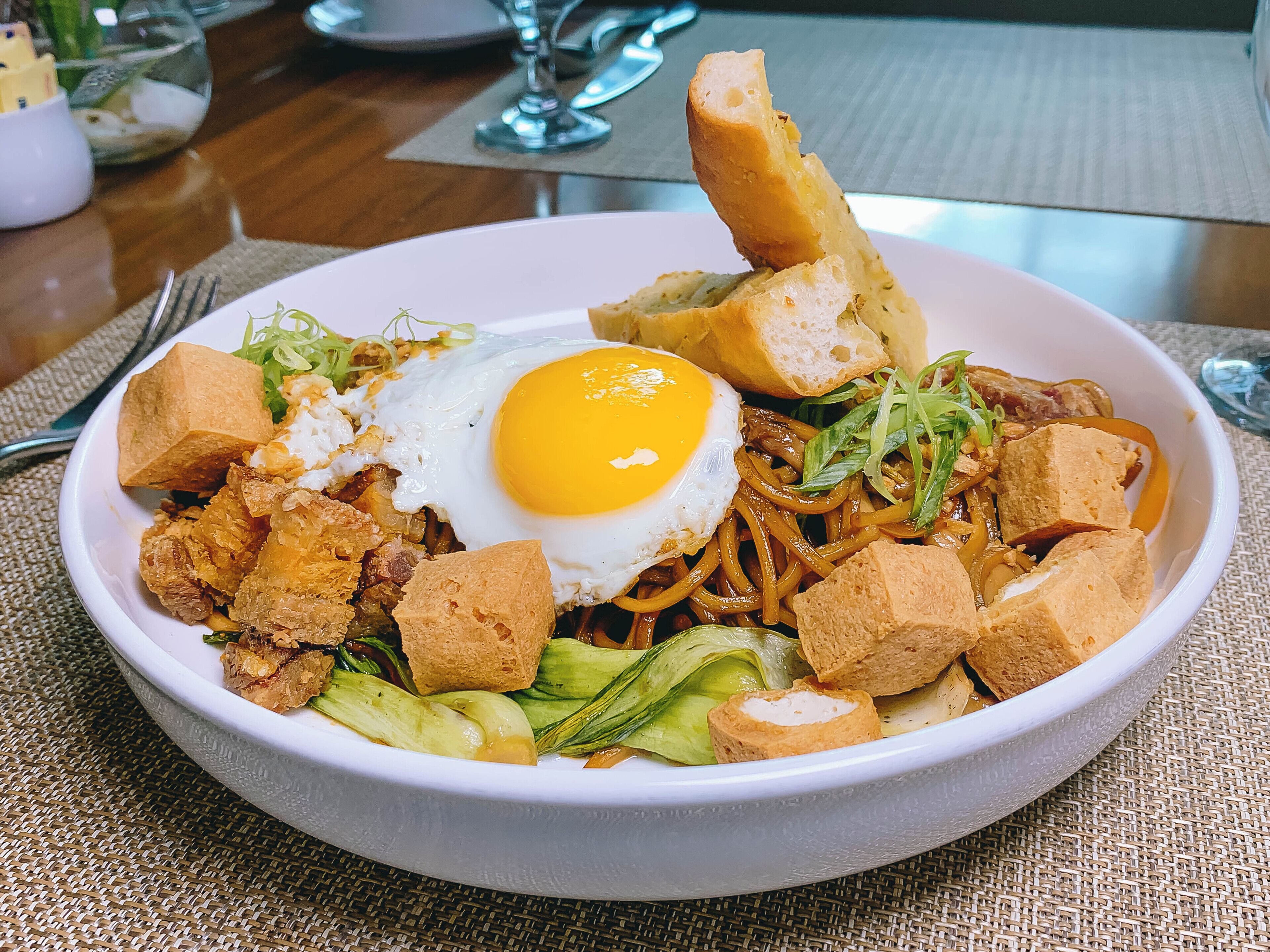 Un plato de comida con un huevo frito, tofu, fideos y verduras.