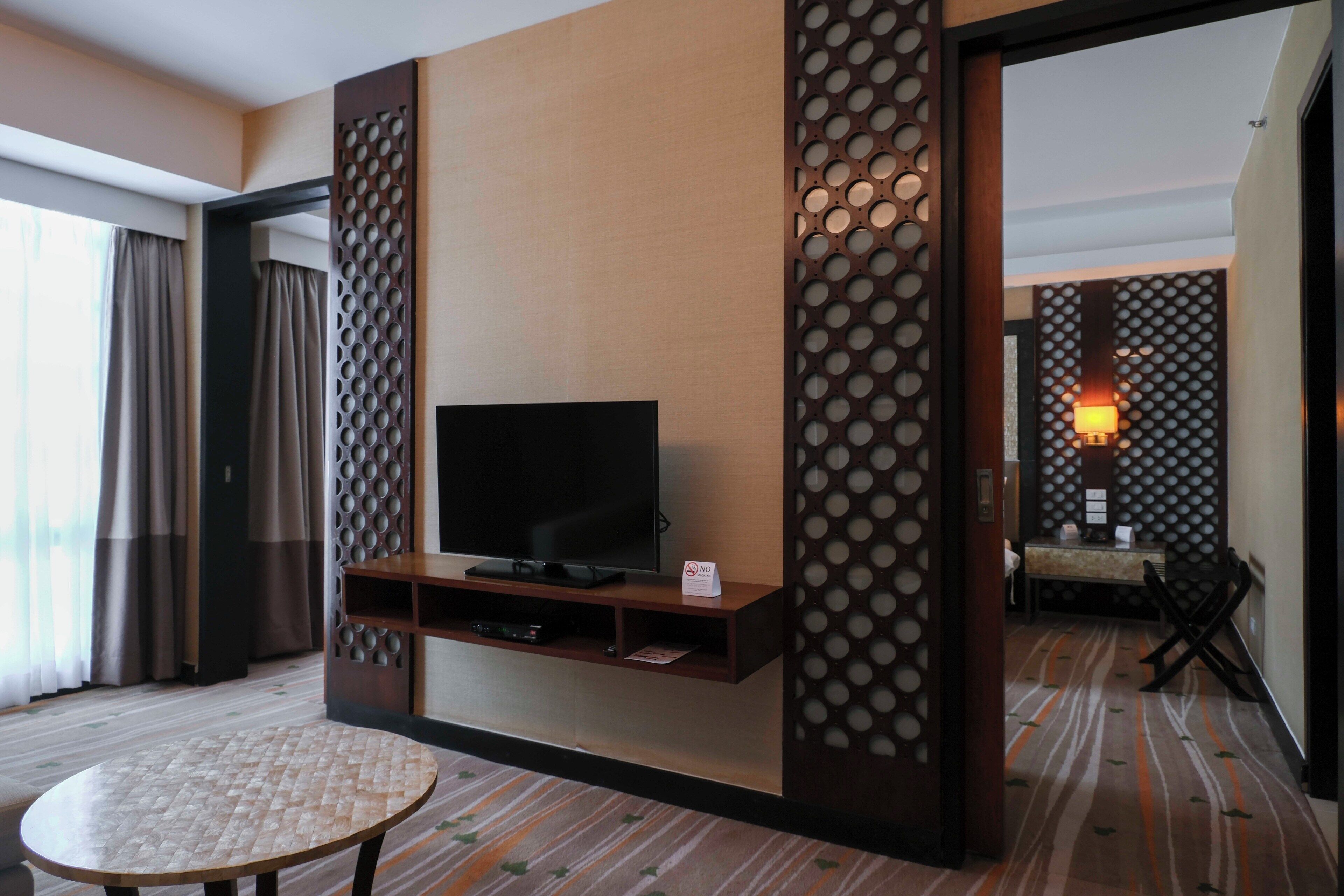 A hotel room with a flat-screen TV on a wooden stand, a small round table, and a decorative wall panel.