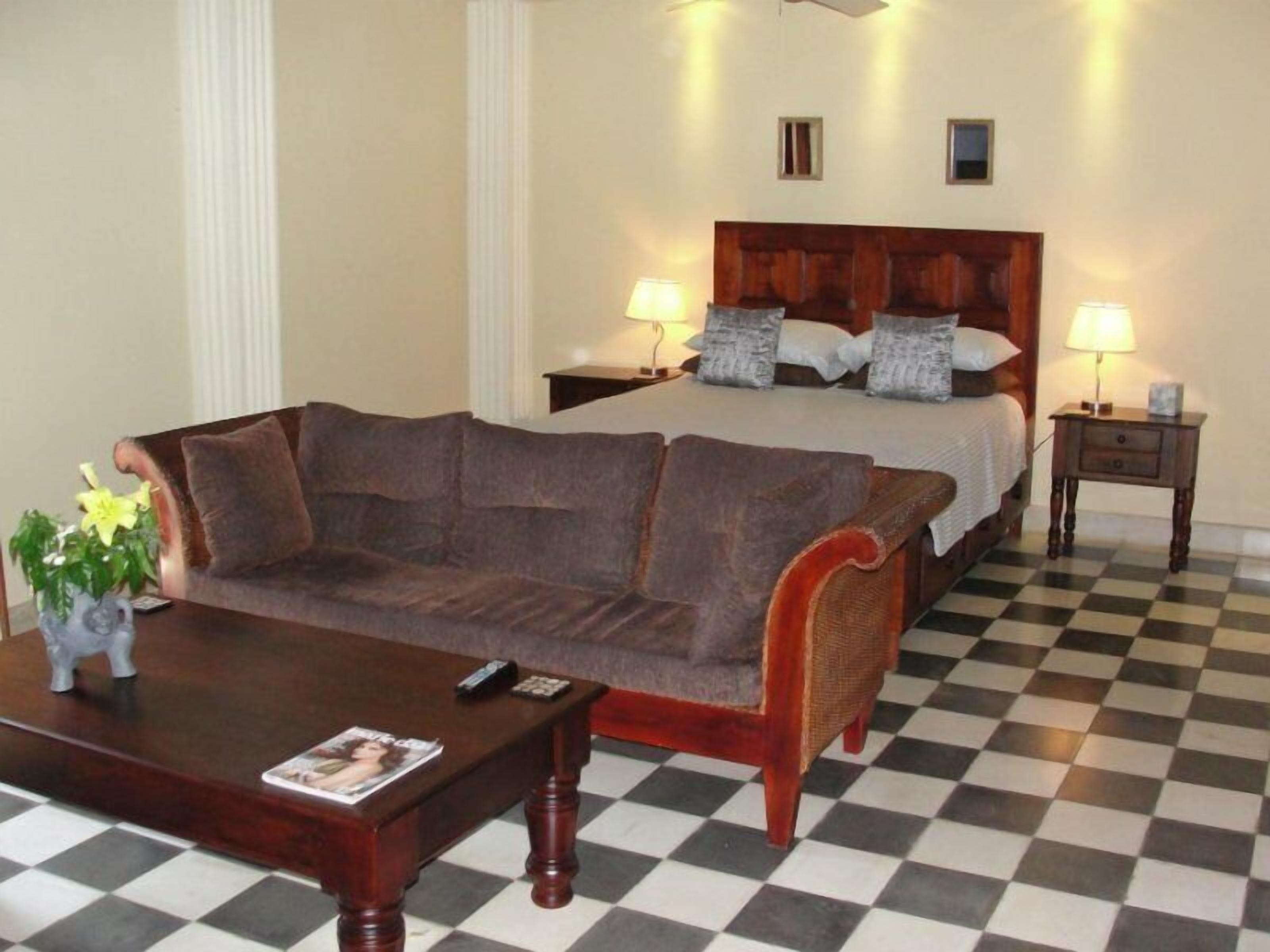 Une chambre d’hôtel avec un sol à carreaux, un canapé marron, un lit en bois avec des oreillers, deux tables de chevet avec des lampes et un vase de fleurs posé sur le canapé.