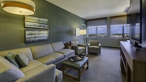 Une chambre d’hôtel moderne dotée d’un canapé d’angle, de fauteuils, d’un bureau avec un ordinateur et de grandes fenêtres offrant une vue sur la ville.