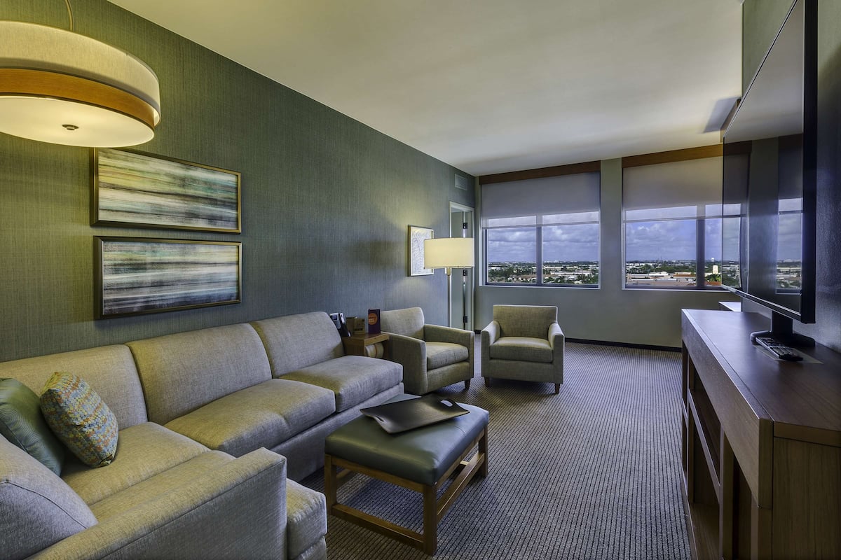 A modern hotel room with a sectional sofa, armchairs, a desk with a computer, and large windows offering a city view.
