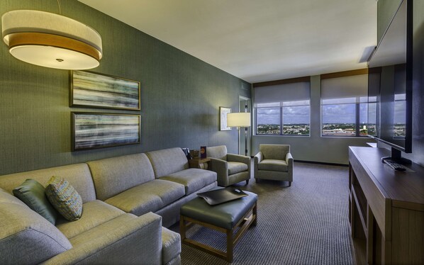 Une chambre d’hôtel moderne dotée d’un canapé d’angle, de fauteuils, d’un bureau avec un ordinateur et de grandes fenêtres offrant une vue sur la ville.