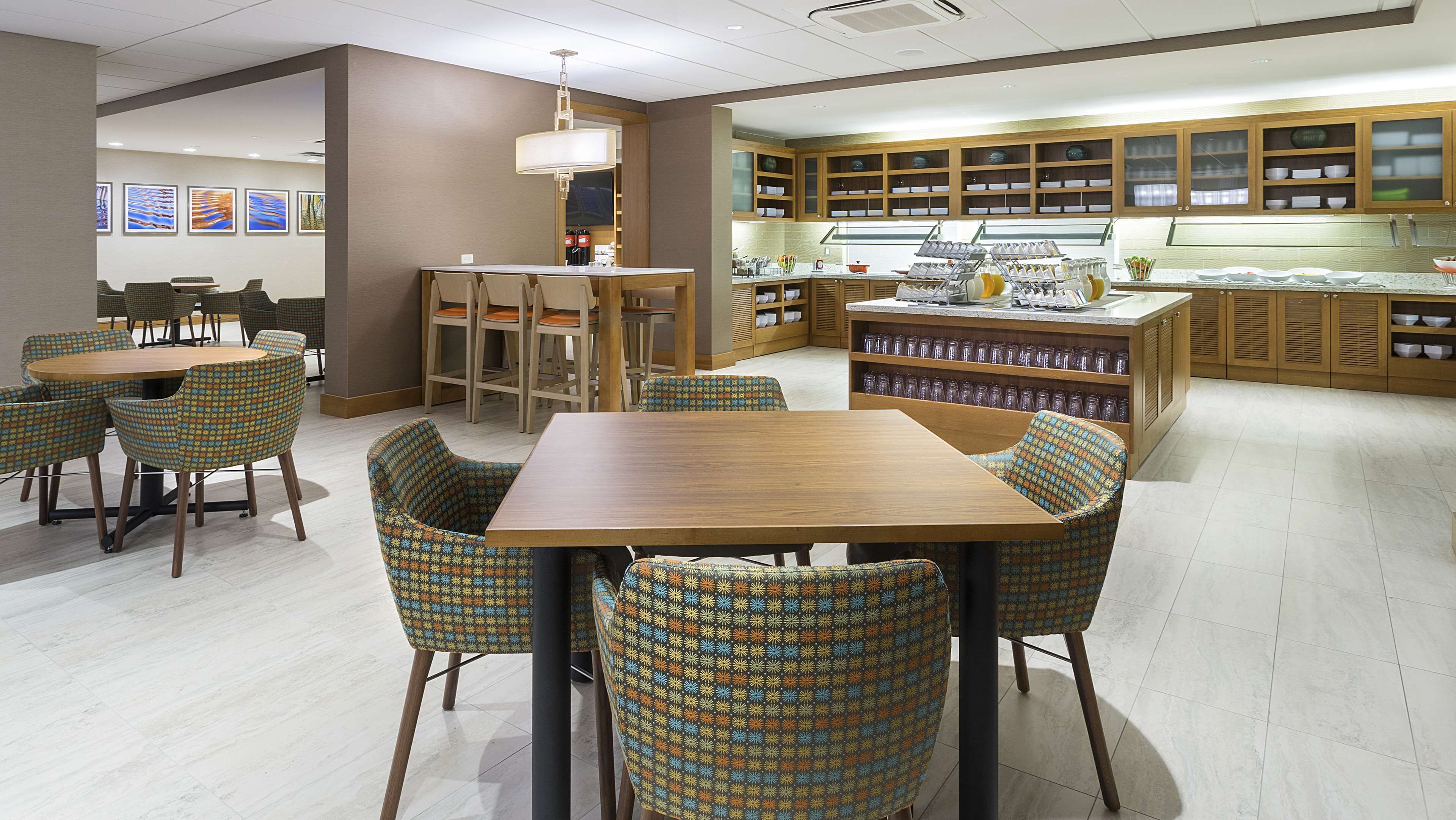 A restaurant with a wooden table and patterned chairs, a kitchen area with shelves of dishes, and a seating area with a large window.