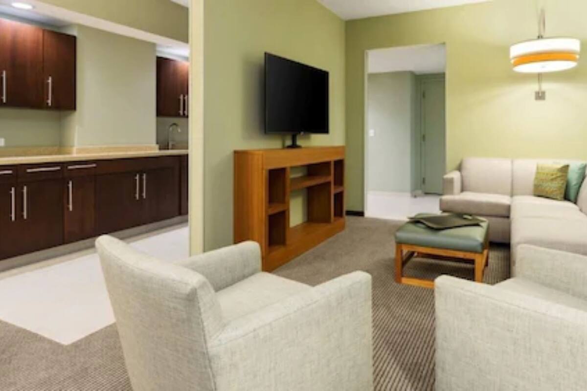 A living room with a sofa, armchairs, a coffee table, a TV, and a kitchen area with cabinets and a countertop.