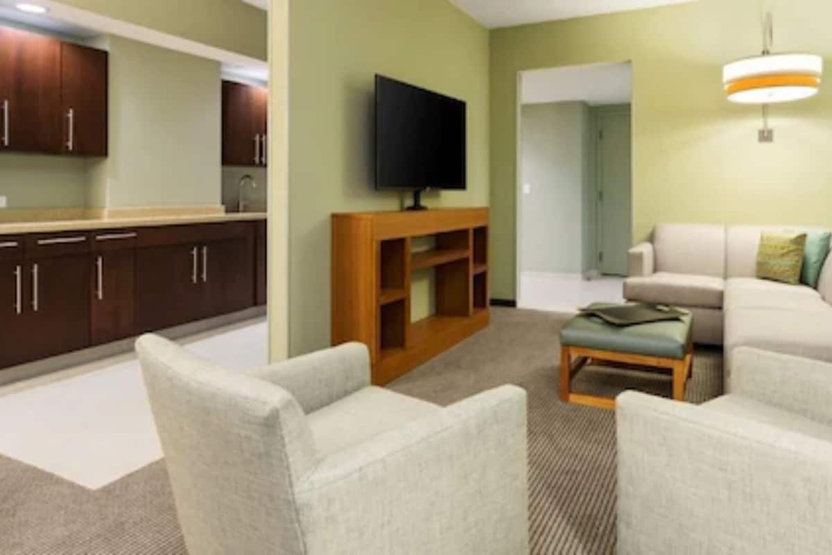 A living room with a sofa, armchairs, a coffee table, a TV, and a kitchen area with cabinets and a countertop.