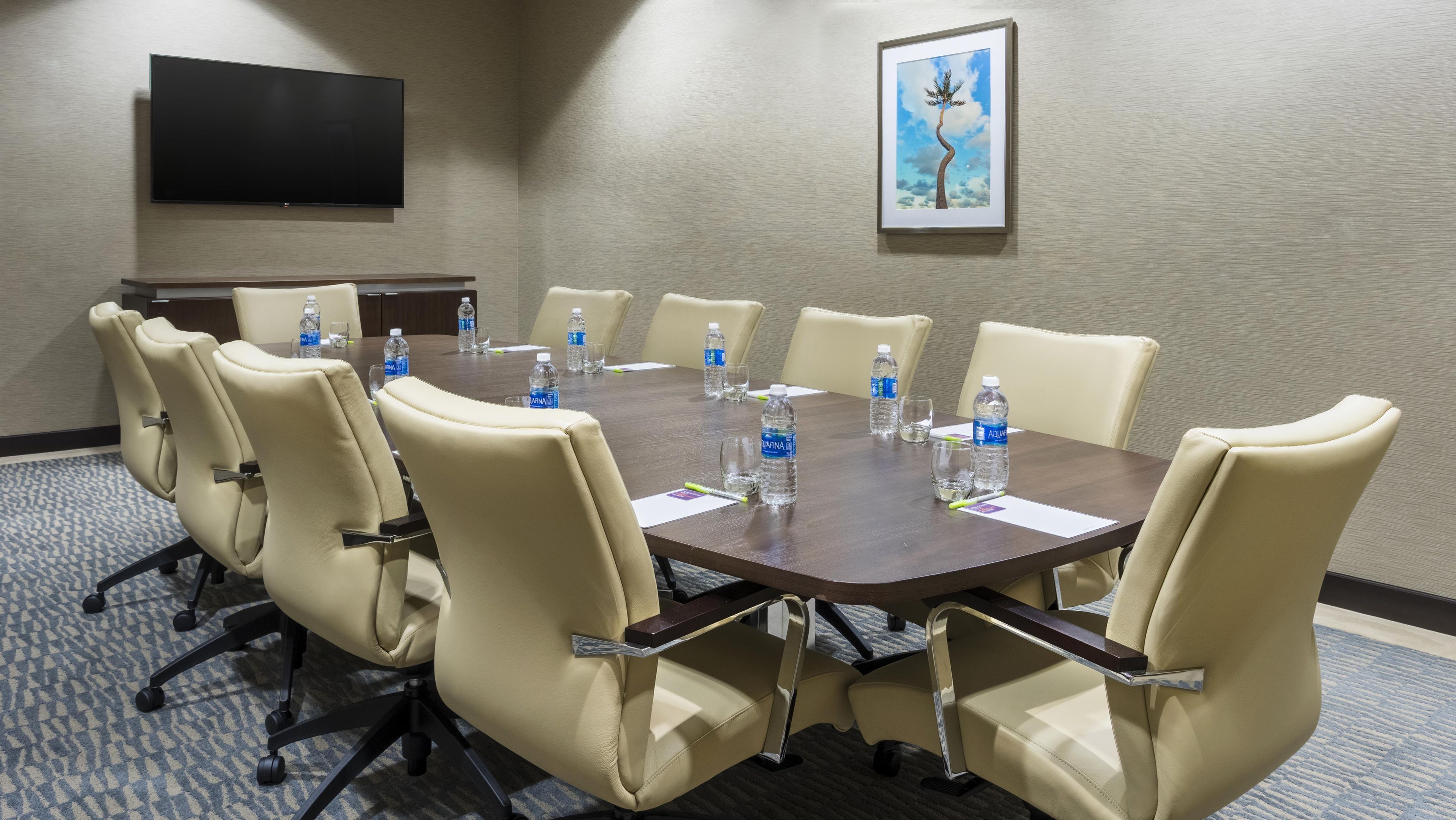Sala de conferência com uma mesa grande, cadeiras, garrafas de água e uma televisão na parede.