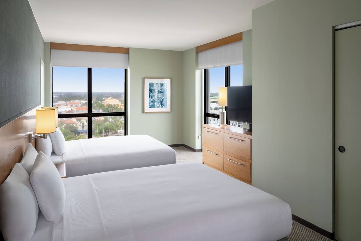 A hotel room with two beds, a TV, a desk, and a window with a city view.