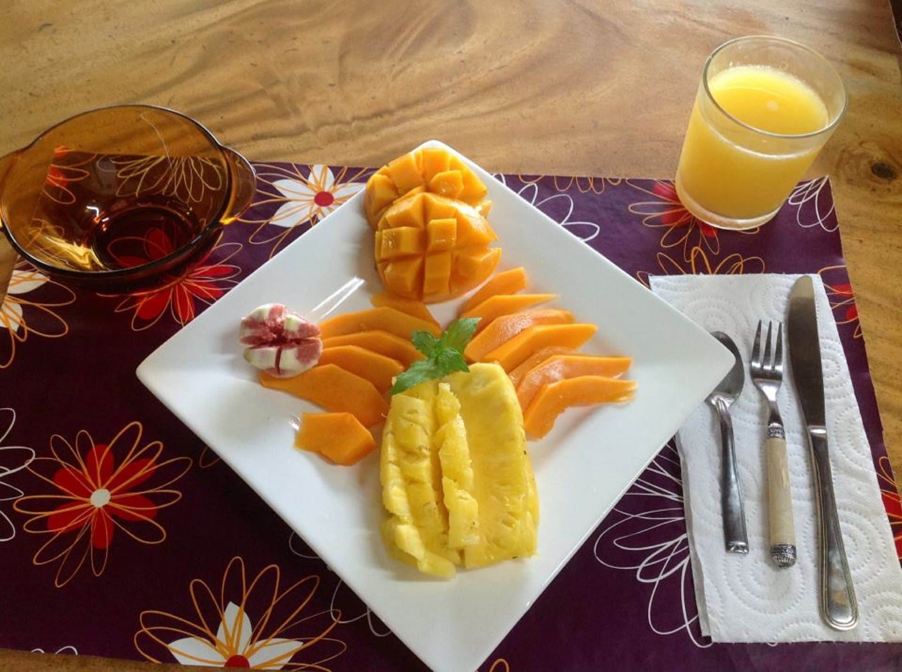 A plate of sliced mango and pineapple, a glass of orange juice, and a bowl of fruit salad on a table with a floral tablecloth.