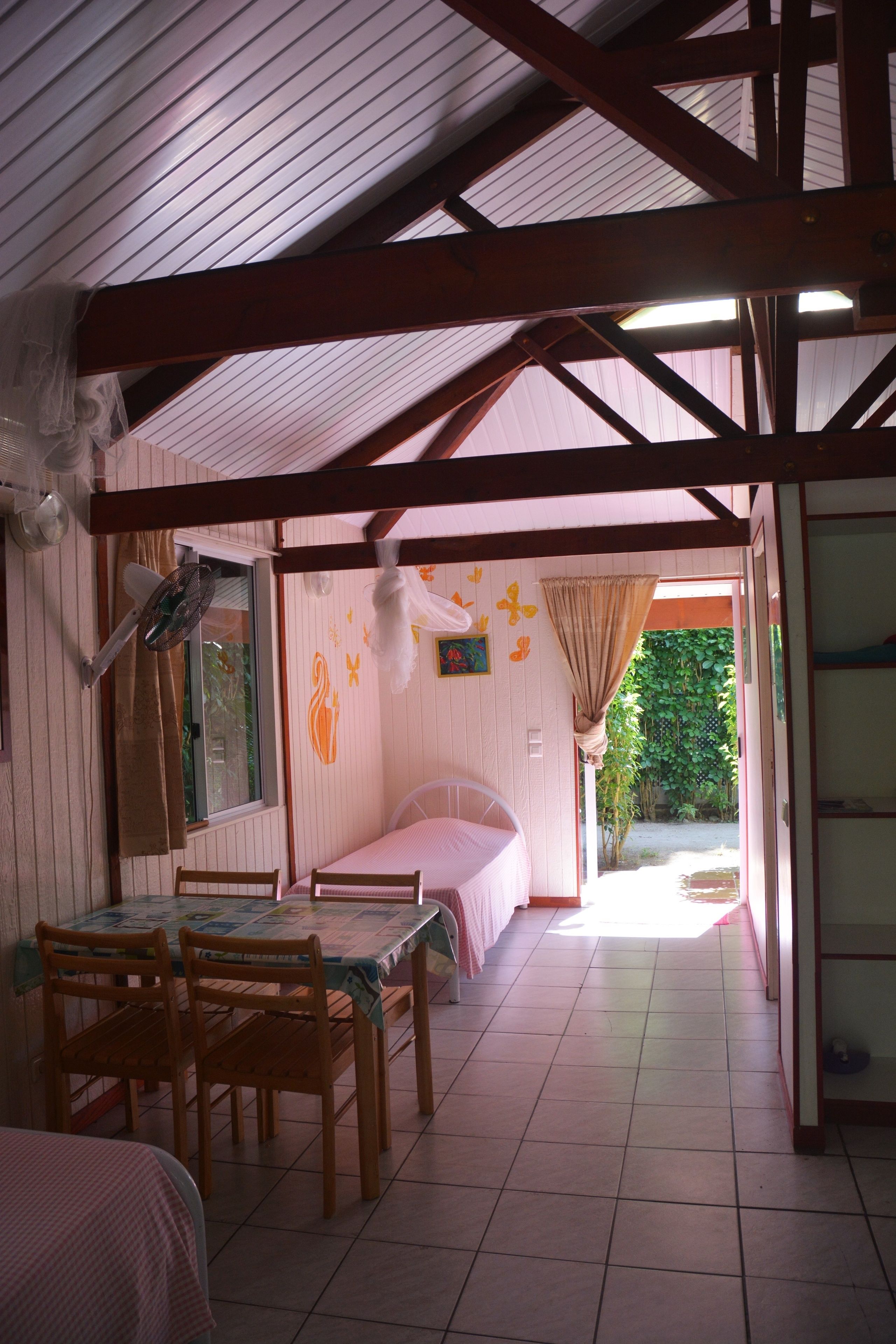 A room with a bed, a table with chairs, a fan, and a window with curtains.