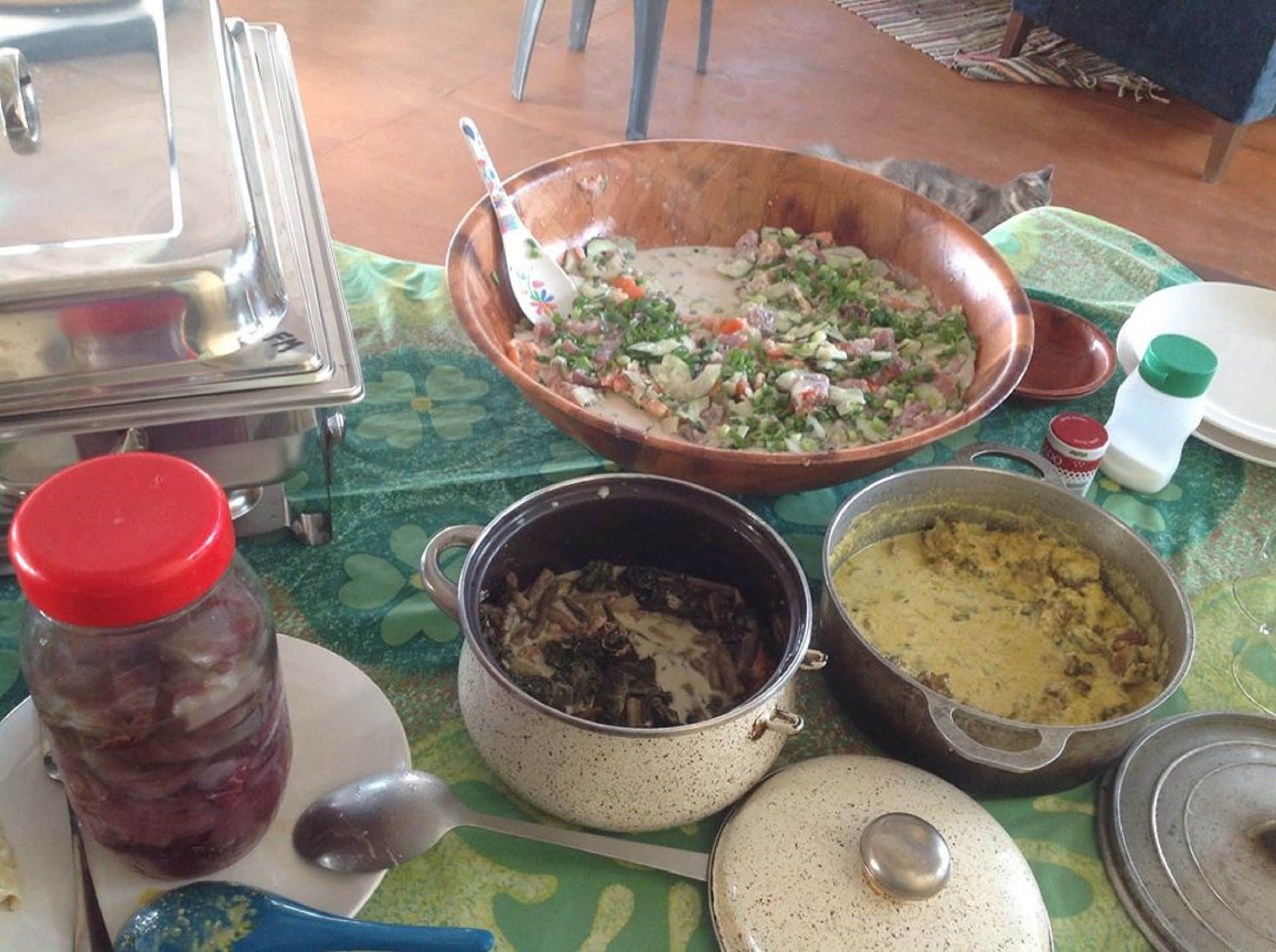 A table with various dishes, including a large bowl of salad, a pot of stew, and a jar of pickles.