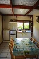 Una cocina compacta con estufa, horno y pileta, sillas de madera alrededor de una mesa con mantel estampado, y una ventana con vista a follaje.