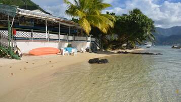 A beachside building with a covered seating area, clear shallow waters, and boats in the background.