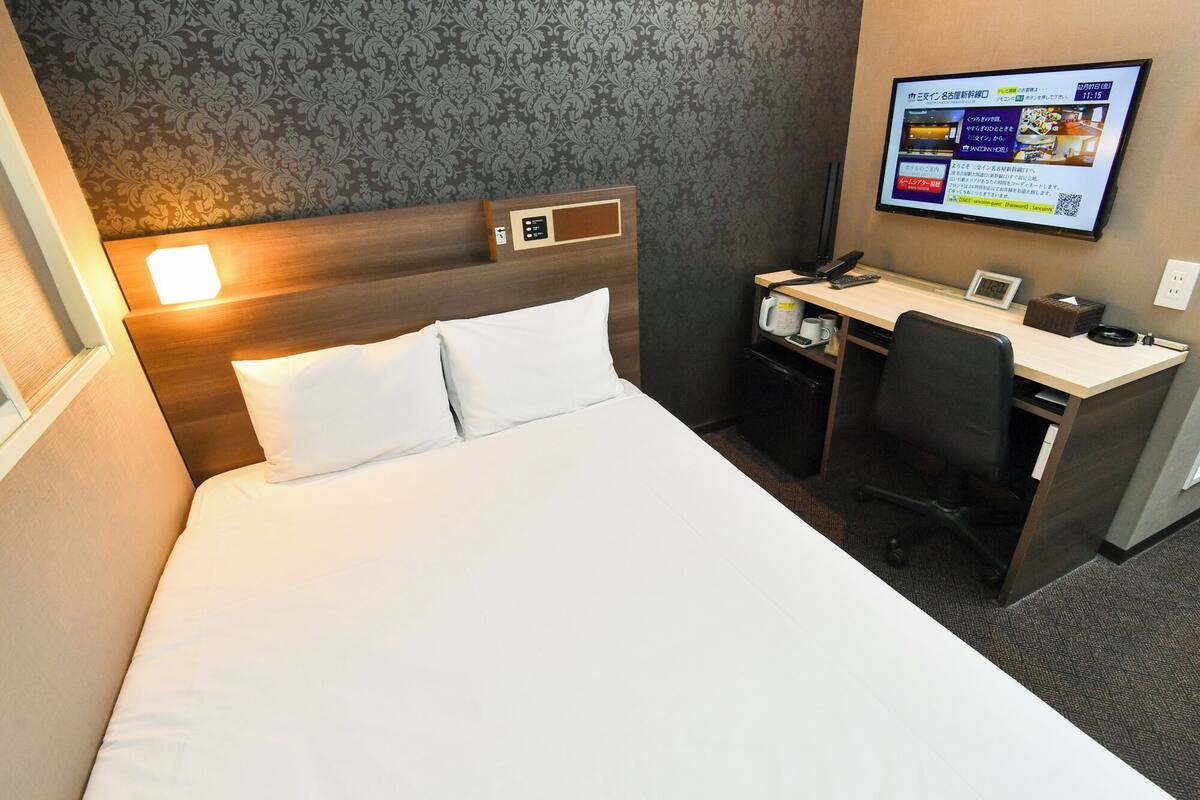 A hotel room with a bed, a desk, a chair, a clock, and a screen displaying information.