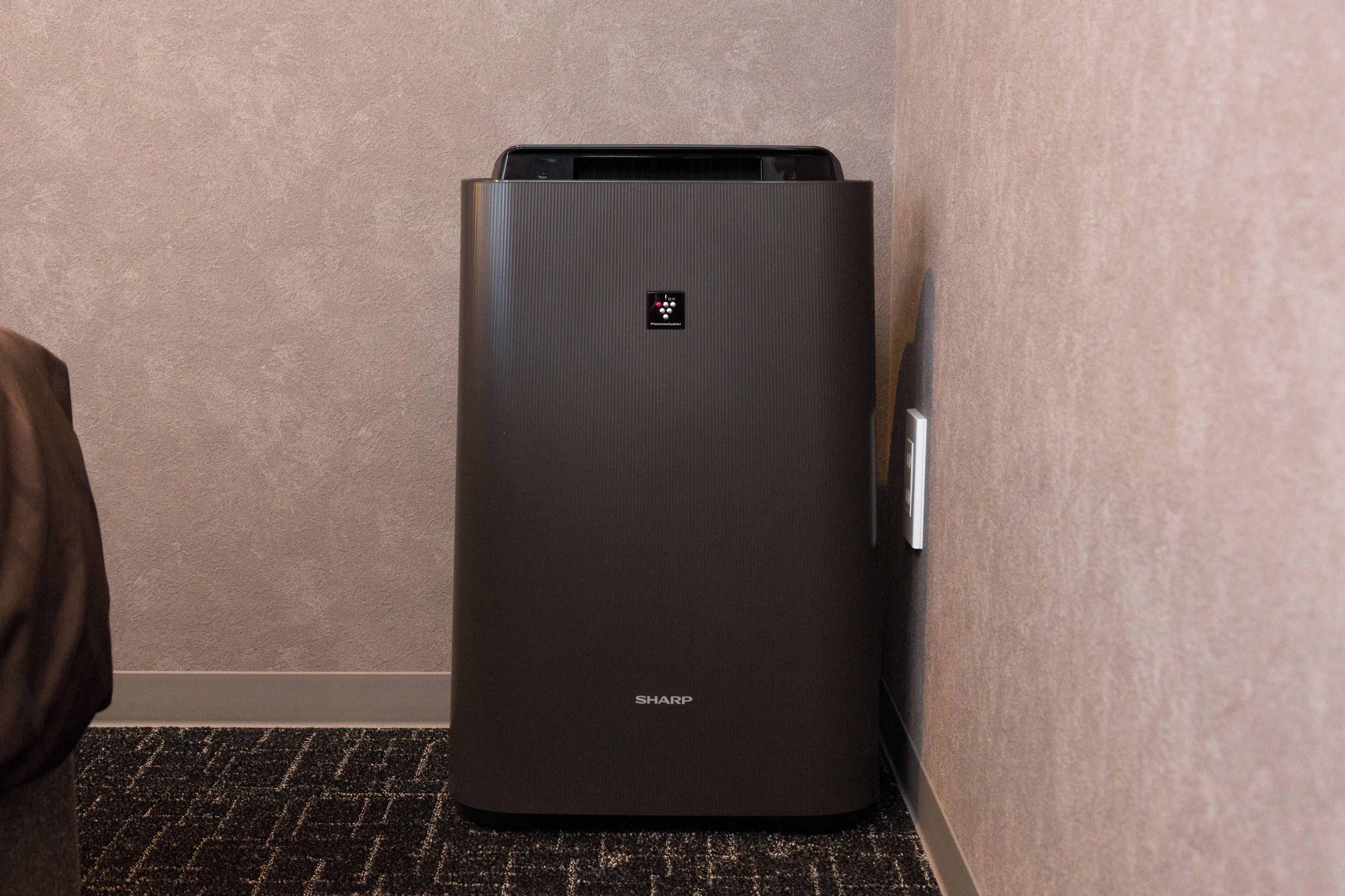 A black, modern, metallic air conditioning unit in a corner of a room with a textured beige wall and a patterned floor.