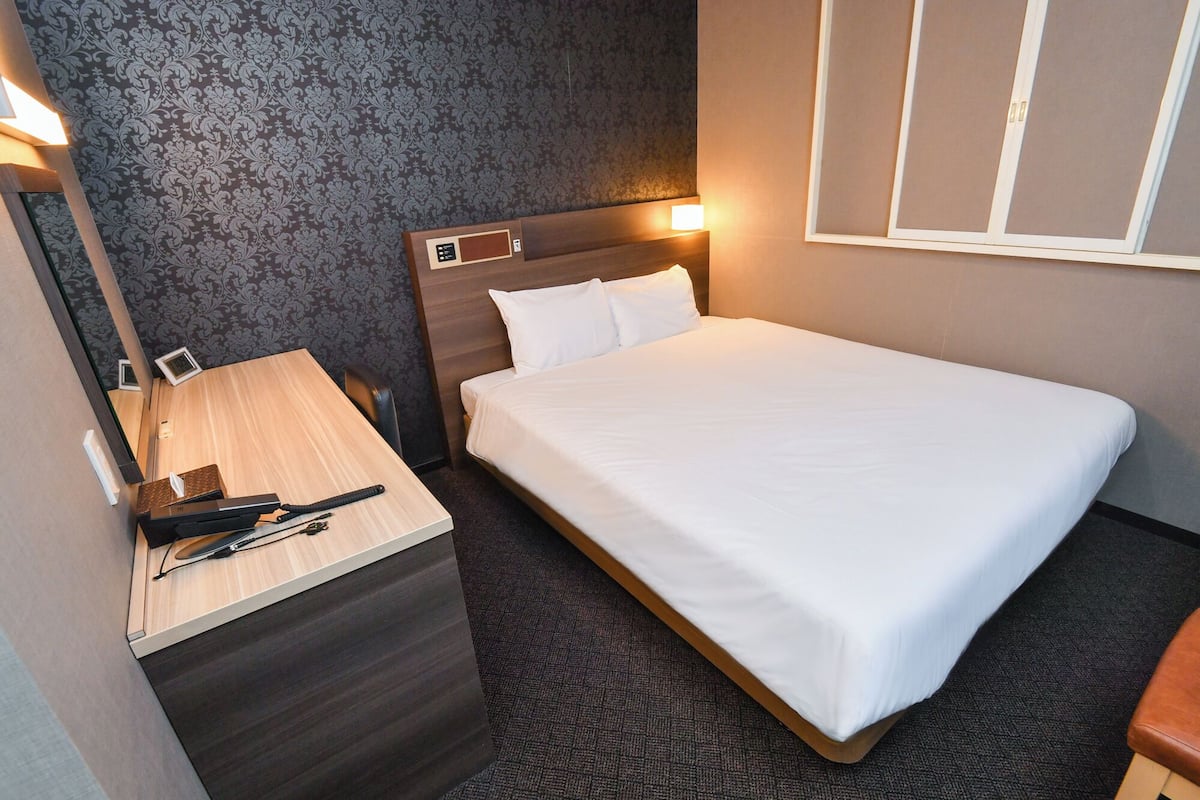 A hotel room with a bed, a desk with a telephone and a notepad, a mirror, and a window with blinds.
