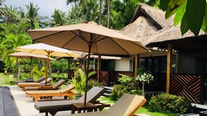 A resort with lounge chairs and umbrellas, surrounded by lush greenery and a thatched-roof building. - Mutiara Bali (Karangasem)