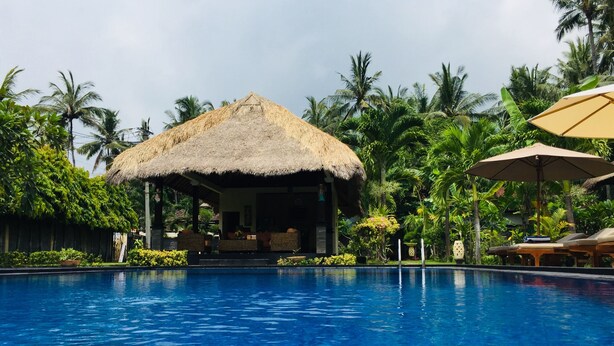 A pool area with a thatched-roof structure, lounge chairs, and palm trees. - Mutiara Bali (Karangasem)