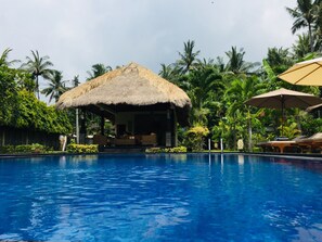A pool area with a thatched-roof structure, lounge chairs, and palm trees. - Mutiara Bali (Karangasem)