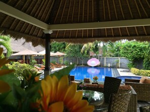 A pool area with lounge chairs and umbrellas, surrounded by lush greenery and a thatched roof structure. - Mutiara Bali (Karangasem)