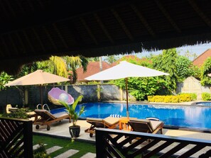 A pool area with lounge chairs and umbrellas, surrounded by greenery and a thatched roof. - Mutiara Bali (Karangasem)