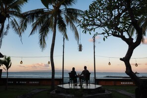 An outdoor seating area with a view of the ocean, surrounded by palm trees and string lights. - Geringsing Bungalow (Karangasem)