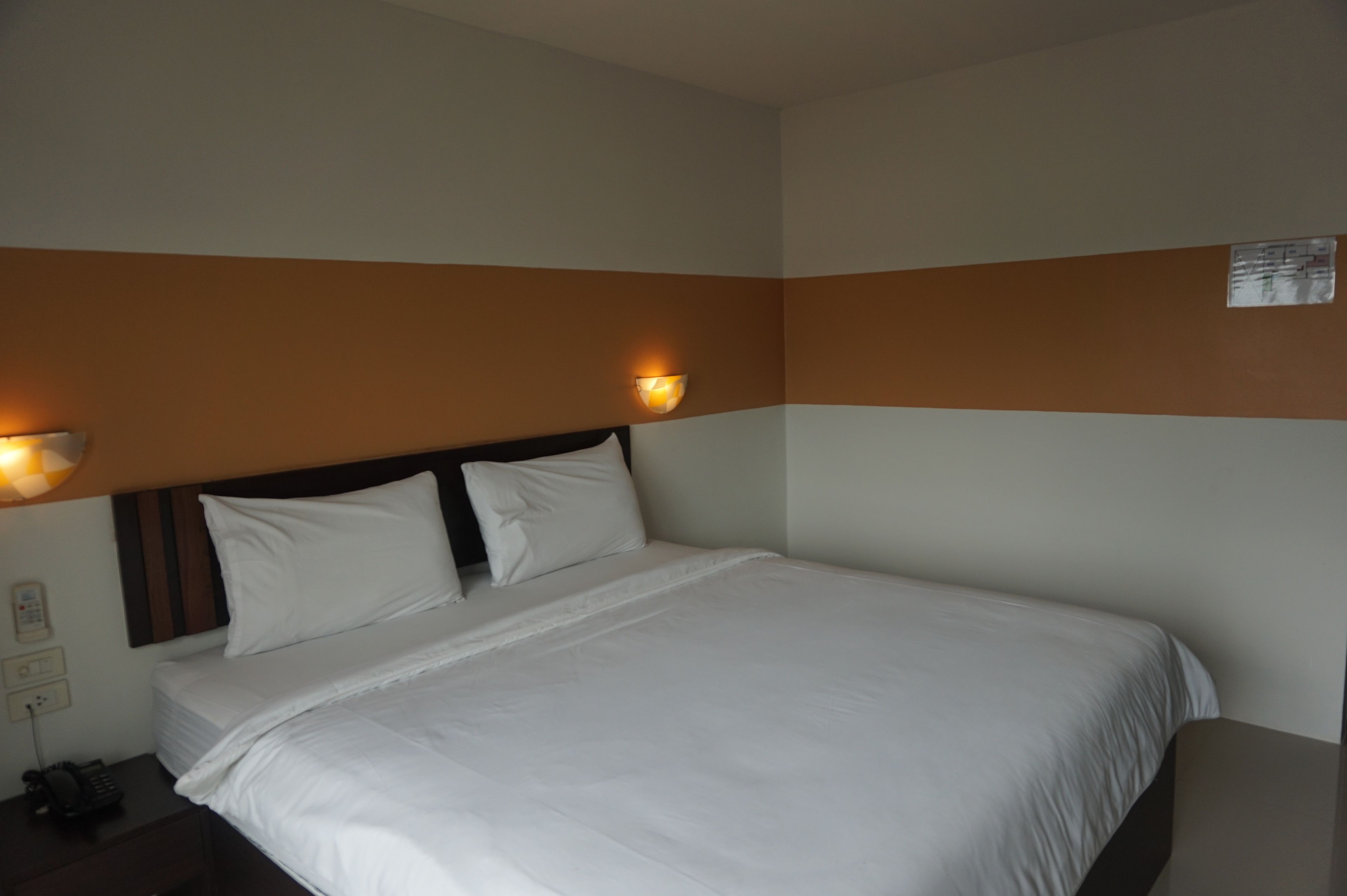 A hotel room with a large bed, two wall-mounted lamps, a nightstand with a phone, and a wall-mounted sign.