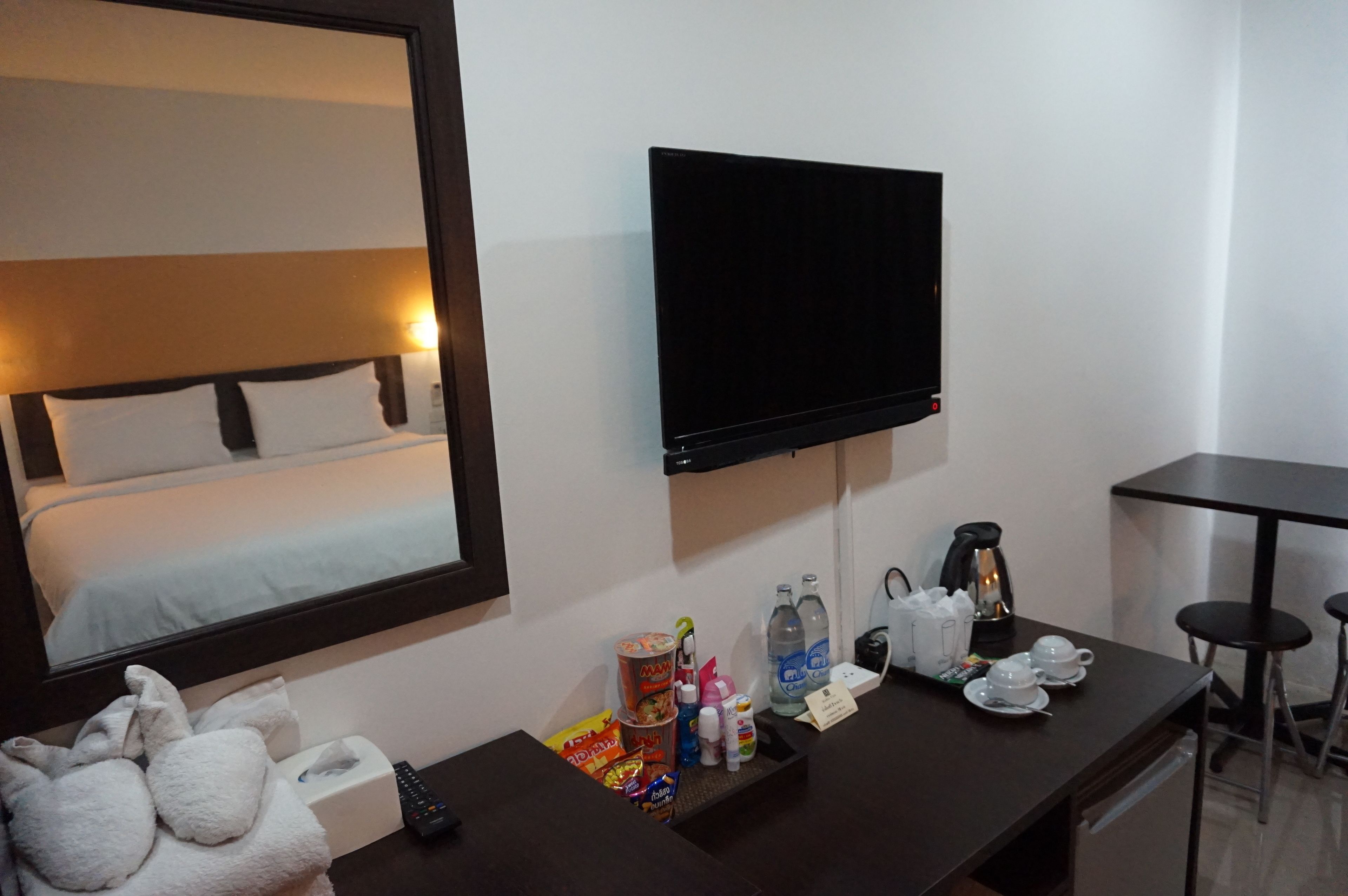 A hotel room with a bed, a TV mounted on the wall, a desk with toiletries and a coffee maker, and a small table with two stools.