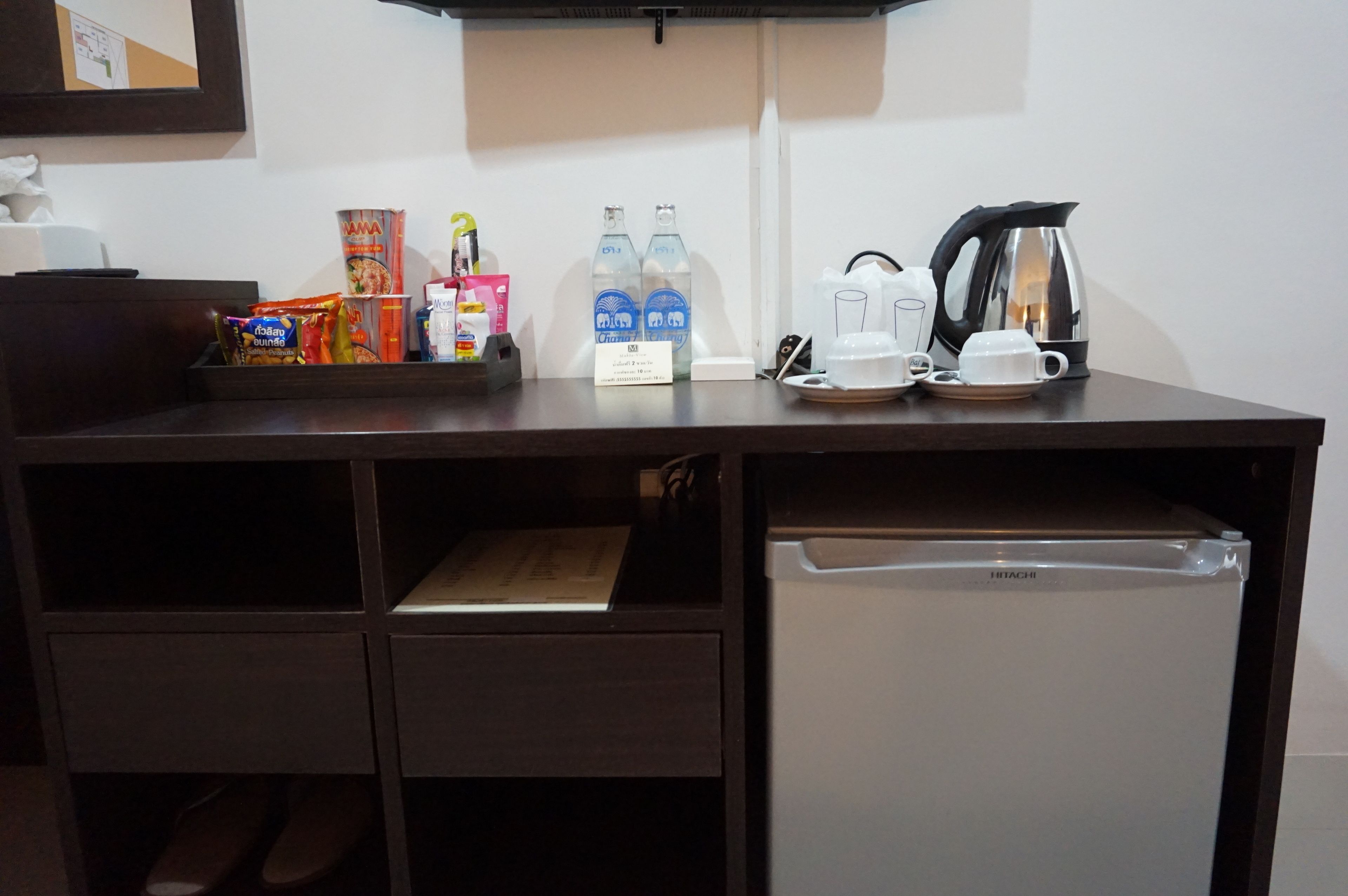 Escritorio de una habitación de hotel con un refrigerador Hitachi, dos botellas de agua, una cafetera y diversos aperitivos.