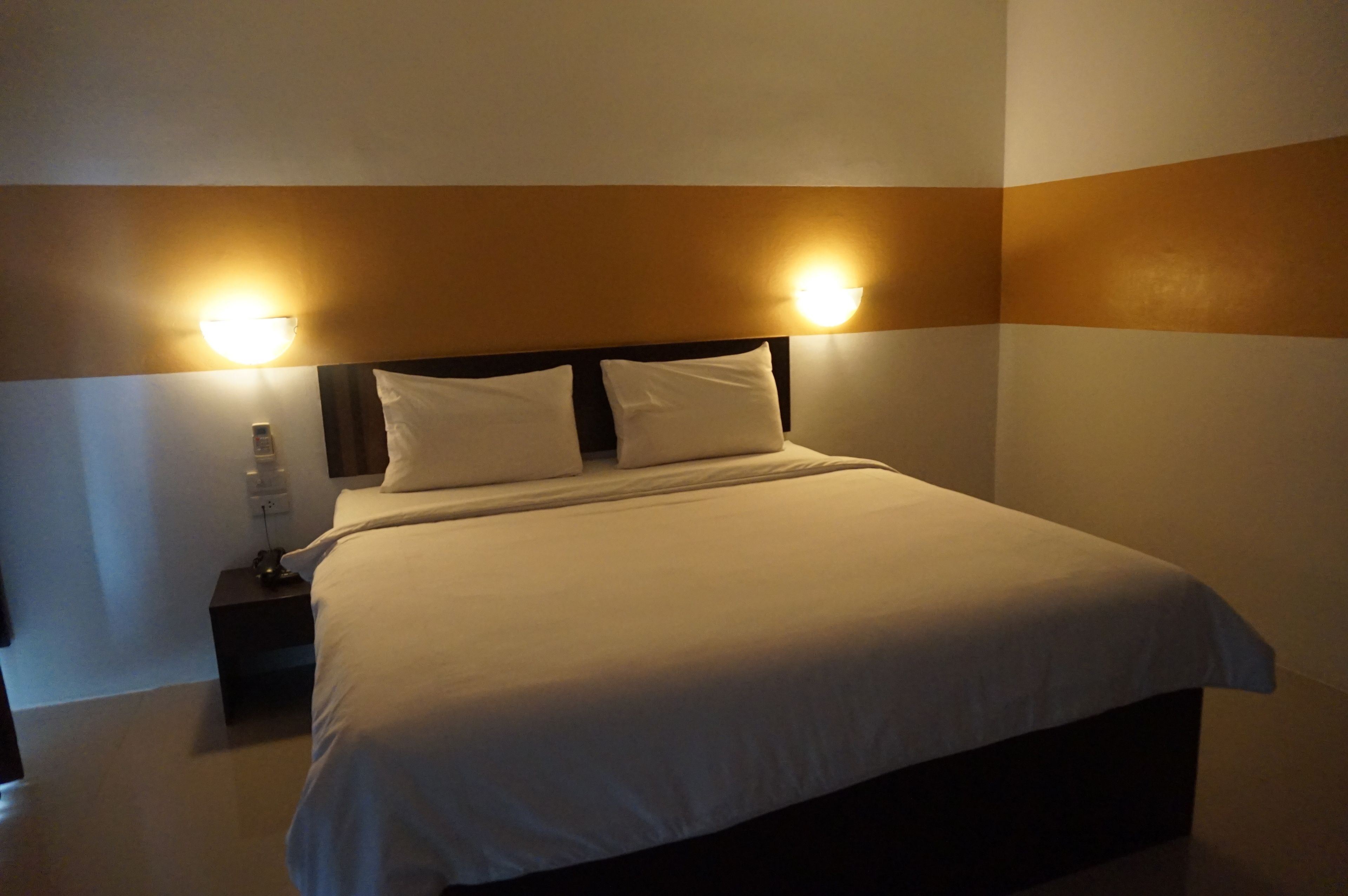 A neatly made bed with white linens, two bedside lamps, and a small nightstand with a bottle and remote control.