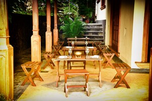 An outdoor dining area with wooden tables and benches, surrounded by plants and a stone wall.