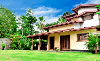 A two-story house with a covered porch, surrounded by lush greenery and palm trees.