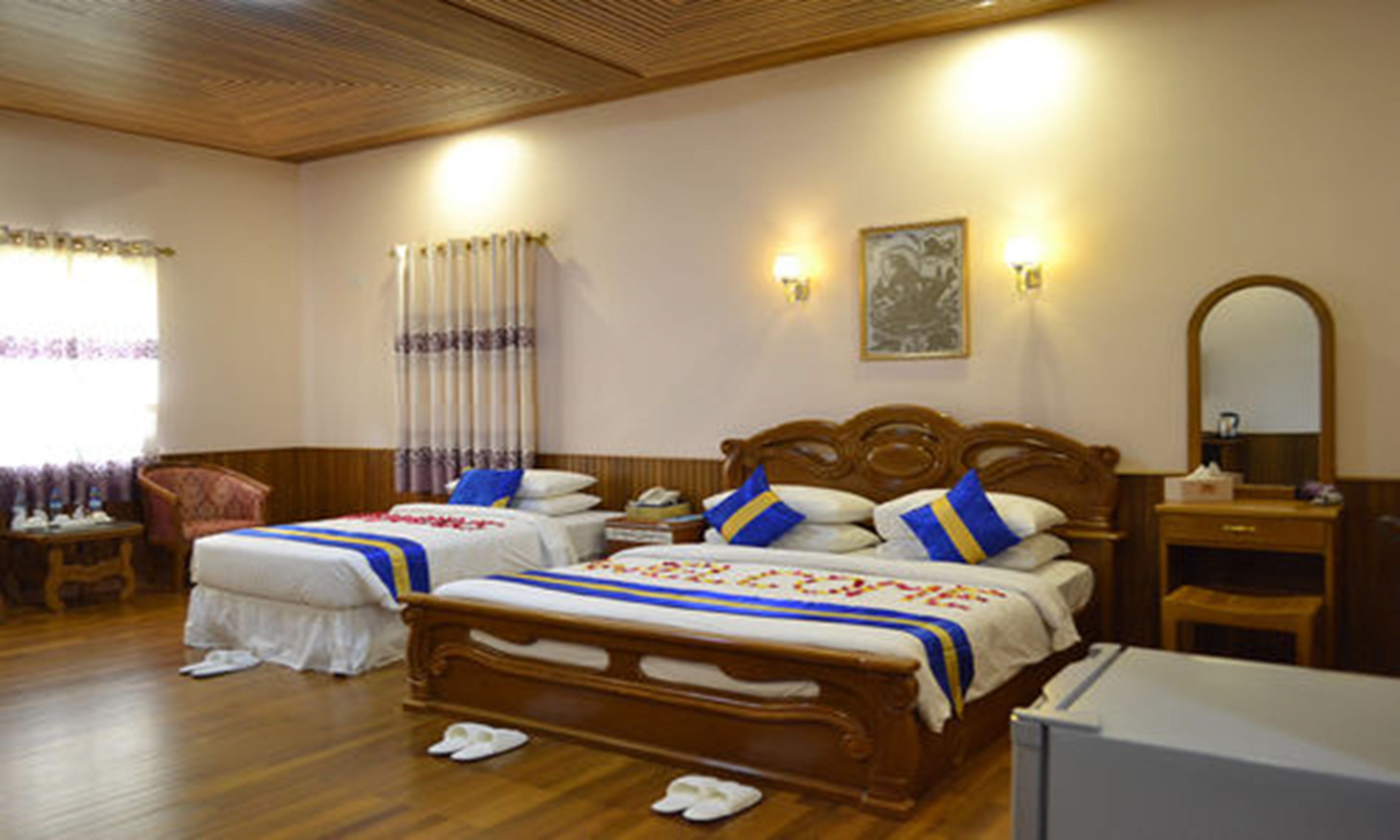 Habitación de hotel con dos camas de madera, una mesita de noche de madera, una silla, un espejo y un ventanal con cortinas.