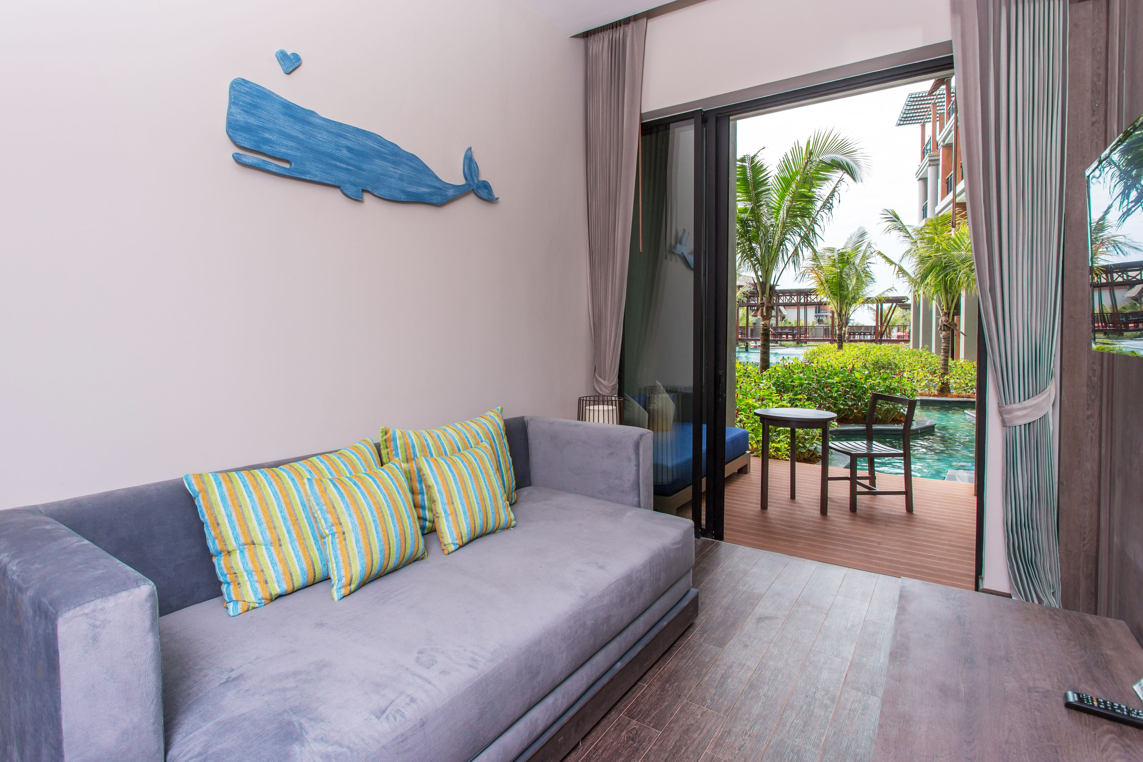 A modern hotel room with a grey sofa, striped pillows, a wooden coffee table, and a view of a pool and greenery outside.