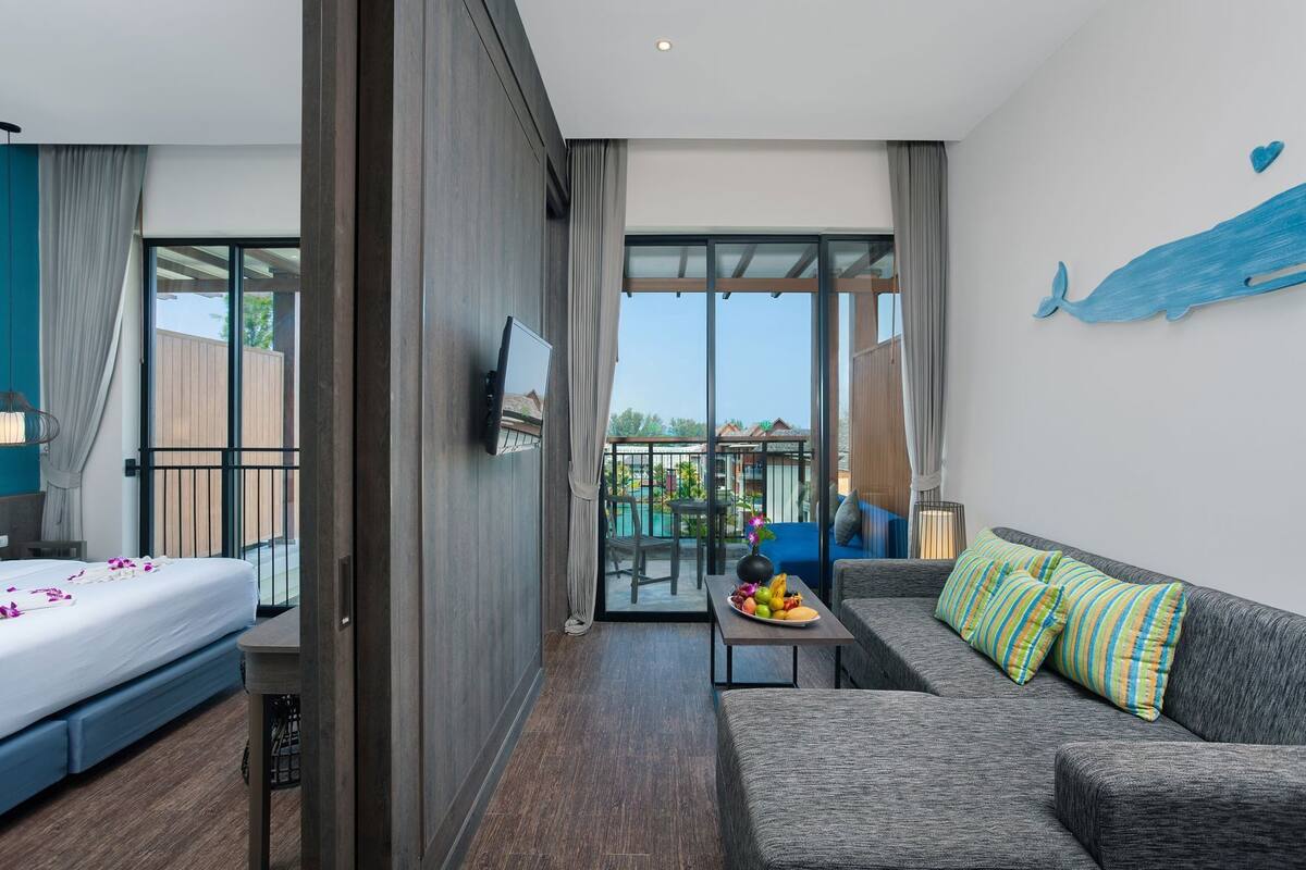 A hotel room with a bed, a sofa, a TV, a balcony with a pool view, and a large window.