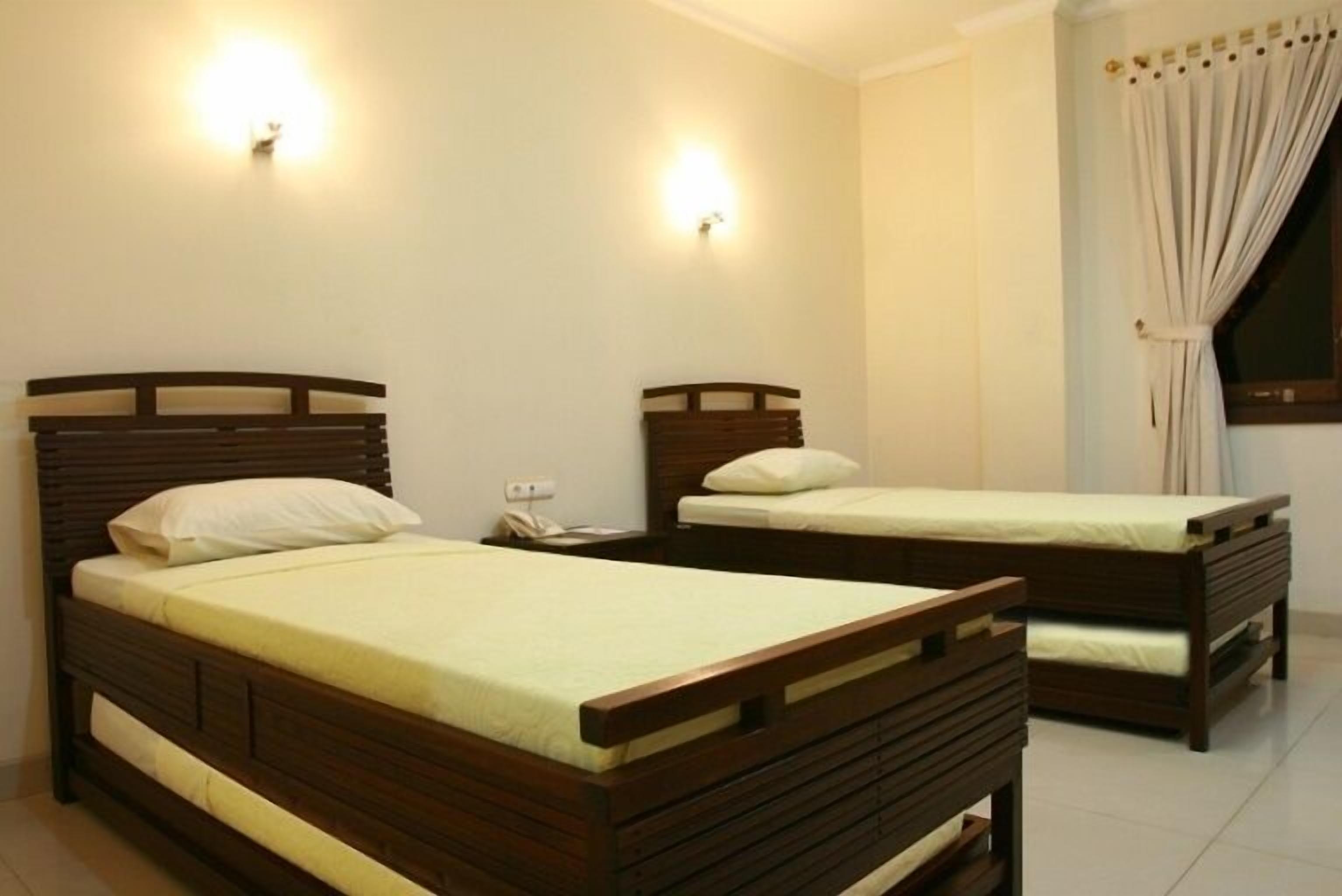 Quarto de hotel com duas camas de madeira, cada uma com travesseiro branco e colchão claro. Há luminárias fixadas na parede acima de cada cama e uma janela com cortinas.