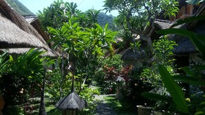 A pathway leading through a lush garden with thatched-roof structures, surrounded by various tropical plants and trees.