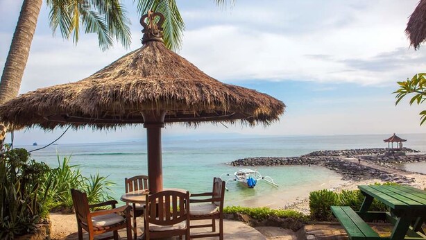 A beachside patio with a thatched umbrella, wooden chairs, and a green picnic table.