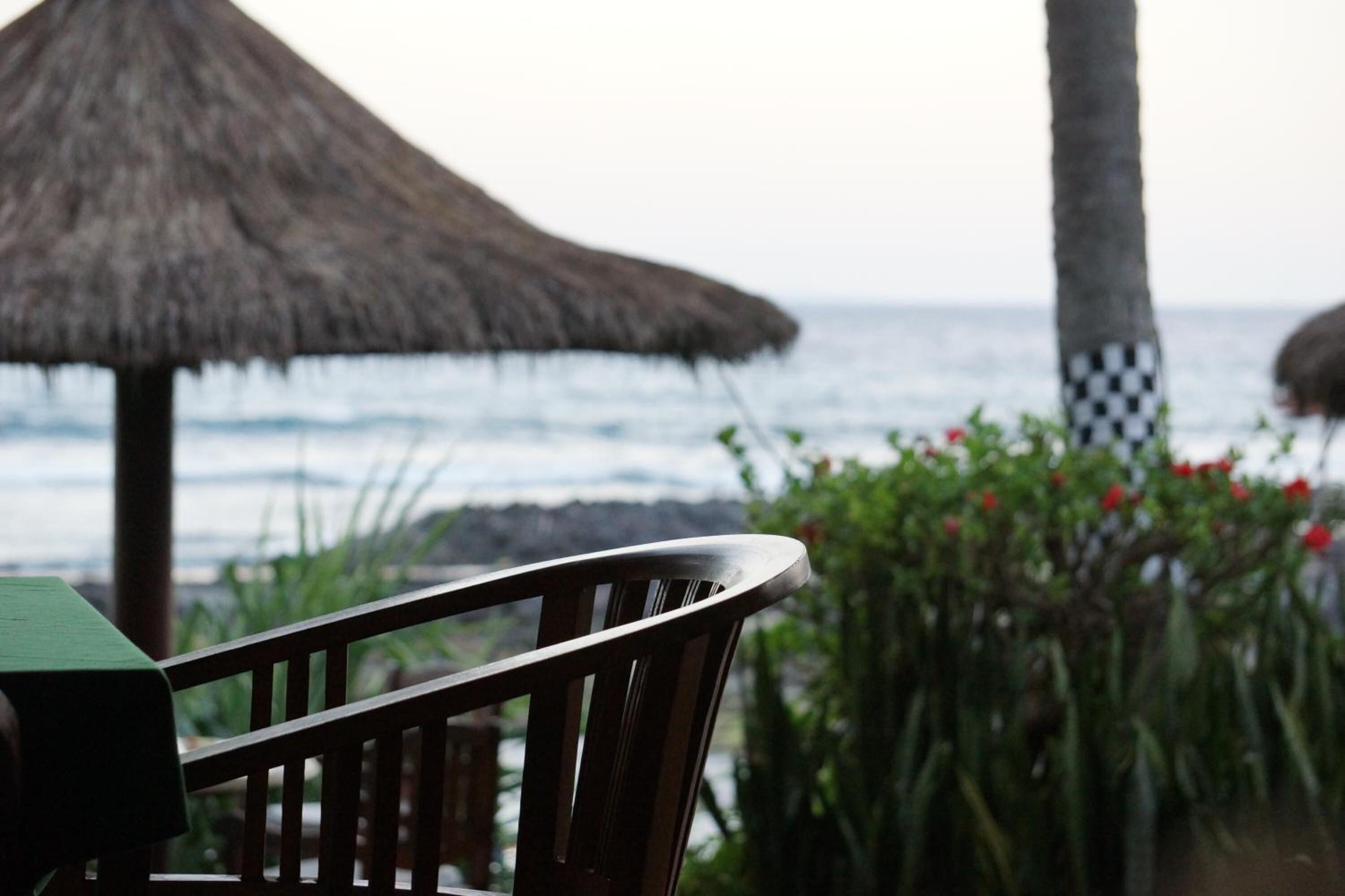 A wooden chair with a green table, a thatched umbrella, and a view of the ocean.